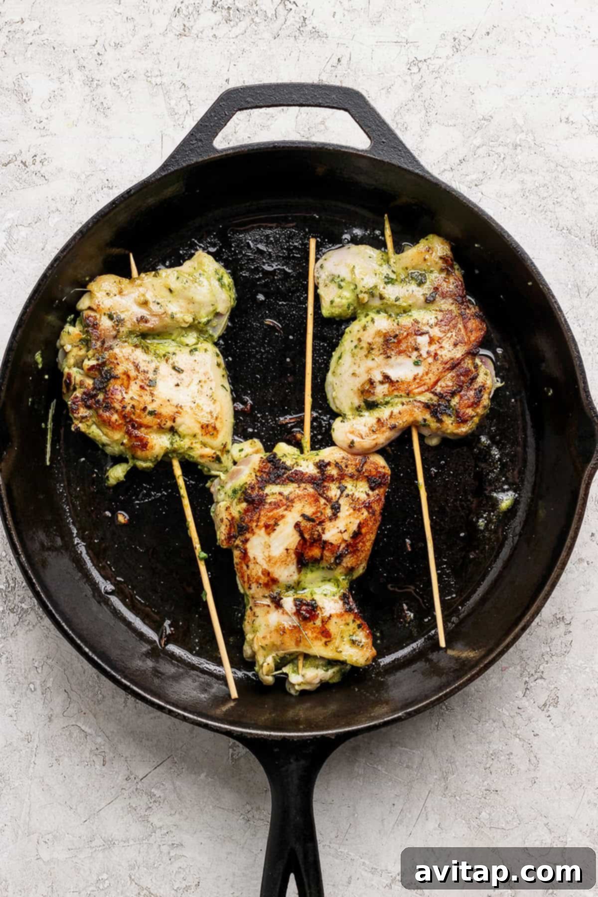 Three grilled, stuffed chicken thighs secured with wooden skewers, sizzling in a black cast iron skillet on a light textured surface.