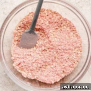 Strawberry Cheesecake Oat Bake 9 A glass bowl filled with pink-tinted oats and a gray spatula resting inside, placed on a light textured surface, showing the dry ingredients mixed with strawberry powder.