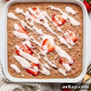 Strawberry Cheesecake Oat Bake 11 A square pan of freshly baked oatmeal, topped with vibrant sliced strawberries and a decadent white icing drizzle, elegantly arranged on a light surface with whole fresh strawberries, a stylish striped towel, and gold utensils nearby, ready to be served.