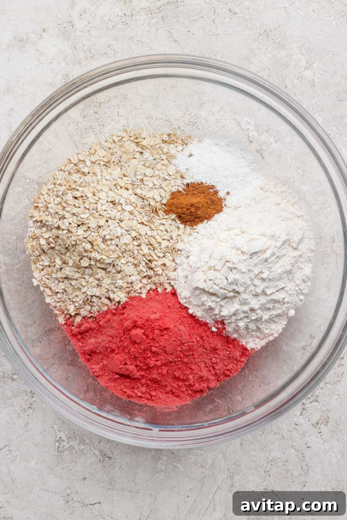 Strawberry Cheesecake Oat Bake 3 A glass bowl containing quick-cooking oats, white flour, pink freeze-dried strawberry powder, cinnamon, baking powder, and baking soda on a light surface, ready to be mixed for baked oatmeal.
