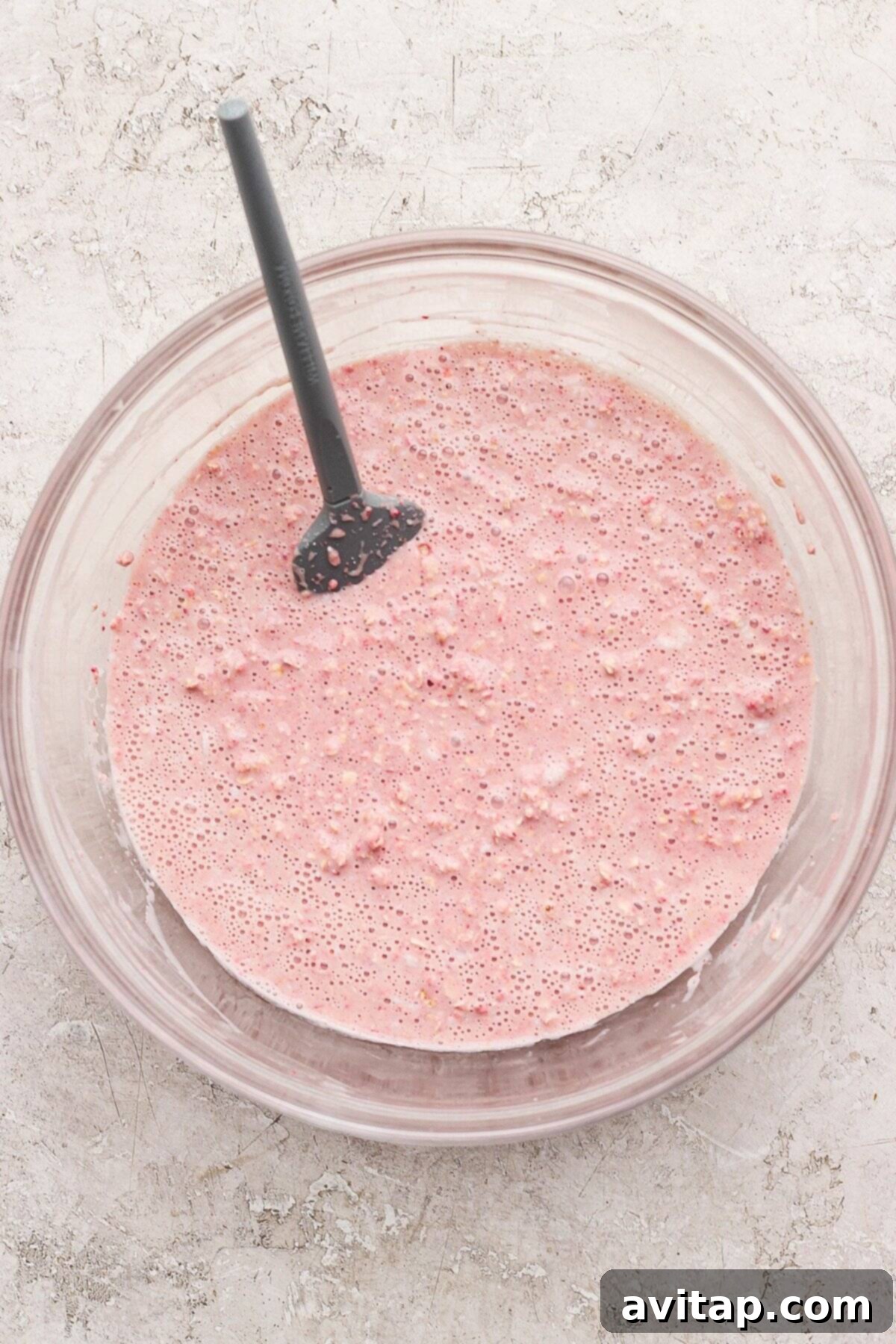 Strawberry Cheesecake Oat Bake 4 A glass bowl filled with a smooth, bubbly pink batter mixture and a black spatula, placed on a light textured surface, showcasing the vibrant color of the strawberry baked oatmeal batter.