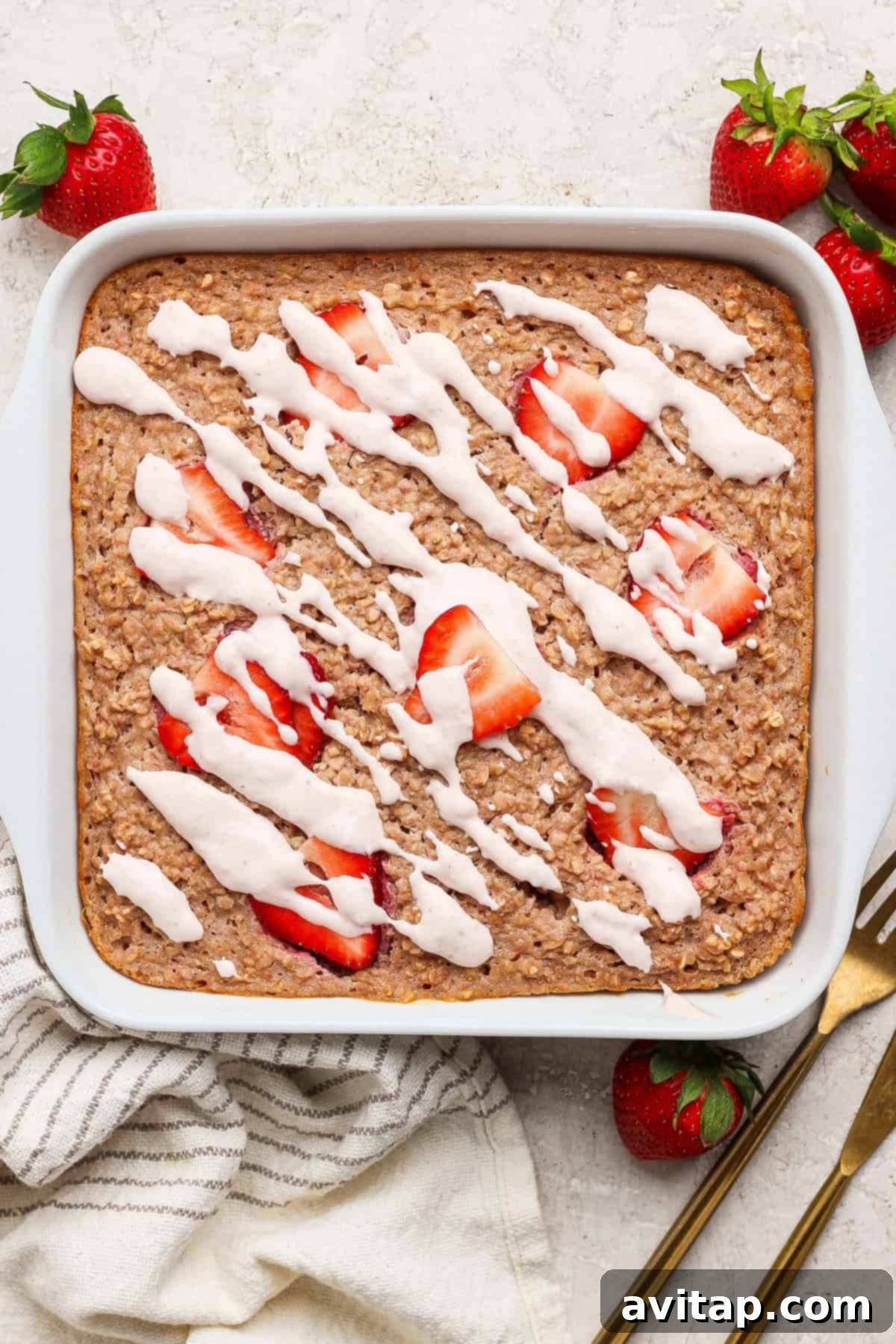 Strawberry Cheesecake Oat Bake 5 A square pan of freshly baked oatmeal, generously topped with vibrant sliced strawberries and a decadent white icing drizzle, elegantly arranged on a light surface with whole fresh strawberries, a stylish striped towel, and gold utensils nearby.