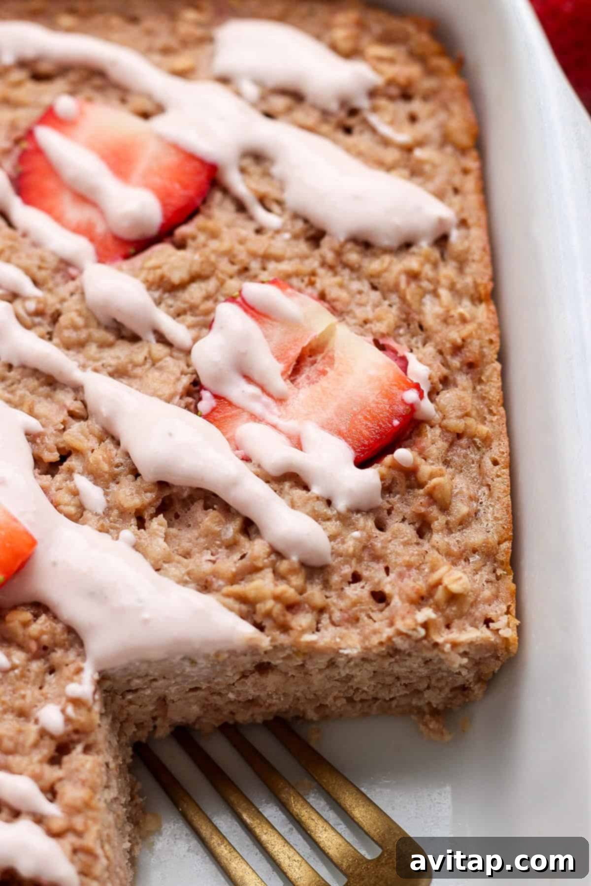 Strawberry Cheesecake Oat Bake 6 Baked oatmeal topped with sliced strawberries and a generous white icing drizzle, still in a square baking dish with a single serving already removed, showcasing a metal spatula nearby.