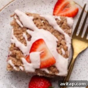 Strawberry Cheesecake Oat Bake 7 A square piece of baked oatmeal cake topped with a generous layer of pink icing and sliced fresh strawberries, served on a speckled plate with a golden fork beside it.