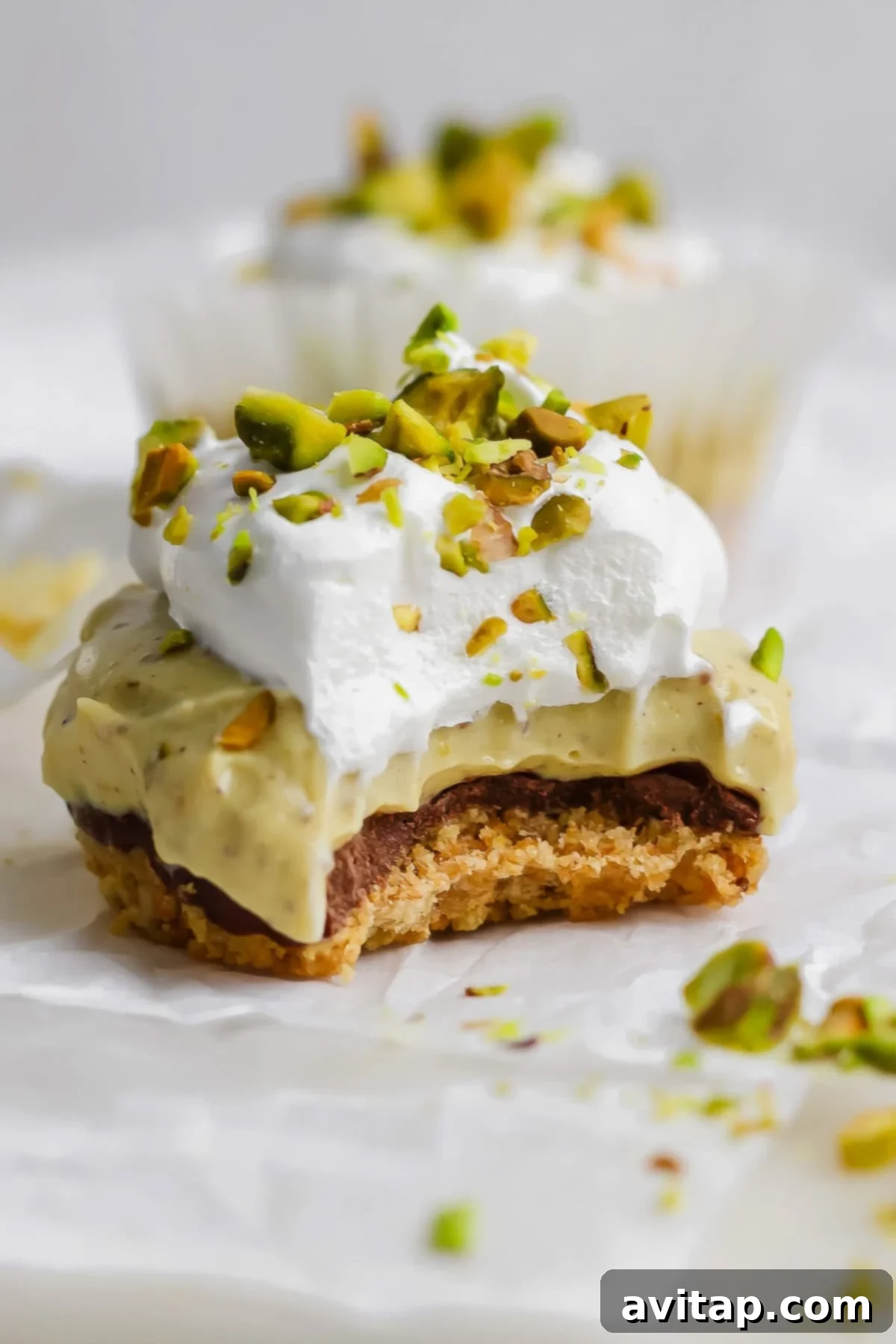 A close-up of a high-protein pistachio cheesecake cup with a crumbly base, creamy green middle layer, fluffy whipped topping, and chopped pistachios, with a bite taken out of it.