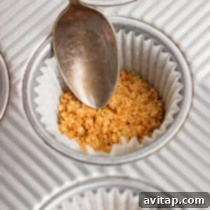 A spoon presses crushed graham cracker crumbs into a paper-lined muffin tin.