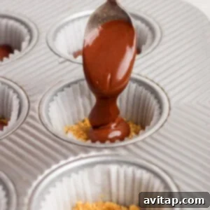 A hand holds a spoon pouring melted chocolate over crushed graham crackers in a paper-lined muffin tin.