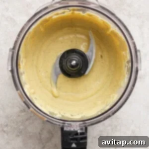A food processor bowl containing a smooth, pale yellow creamy mixture with the blade visible in the center, set on a light textured surface.