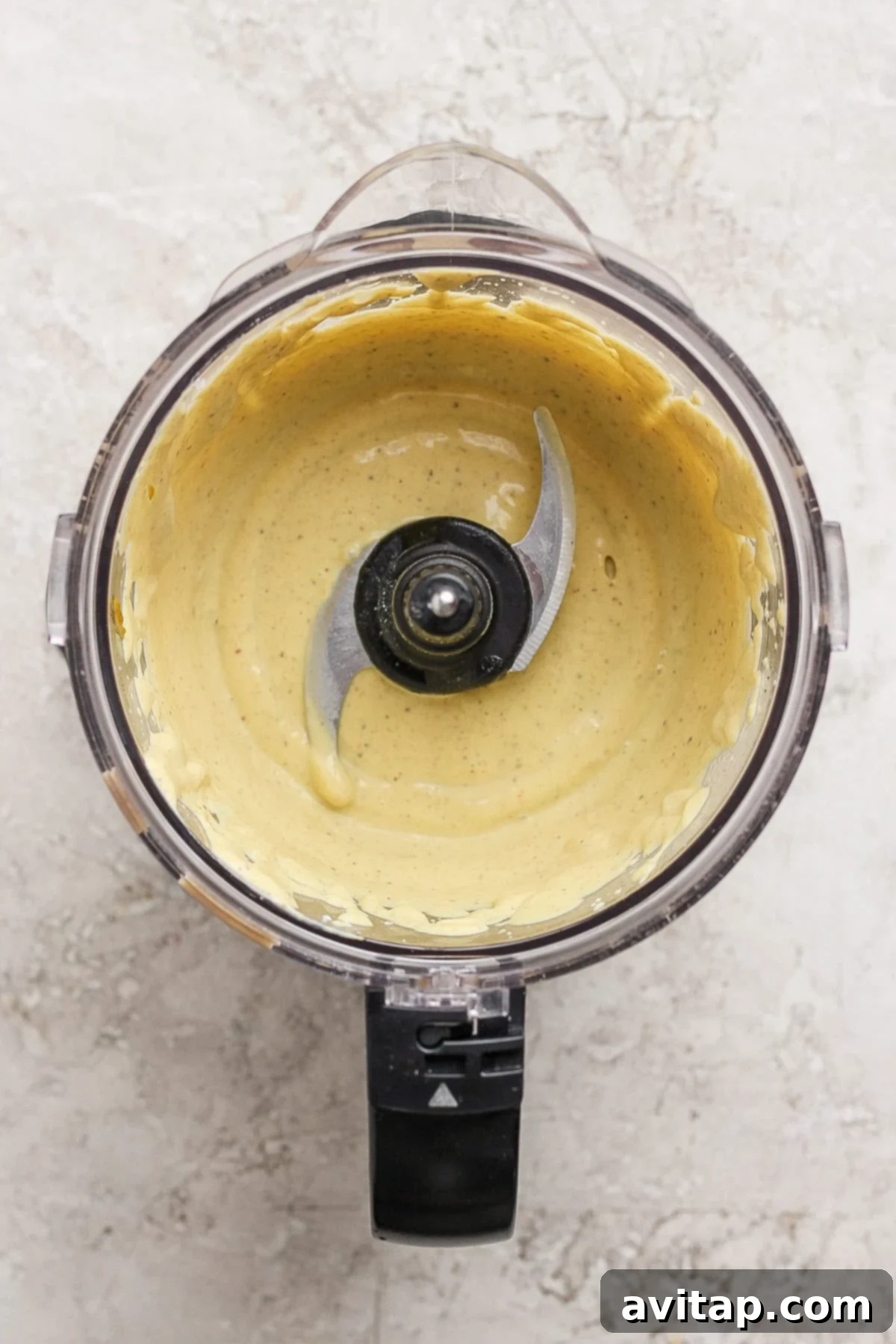 A food processor bowl containing a smooth, pale yellow creamy cottage cheese mixture, with the blade visible in the center, set on a light textured surface.