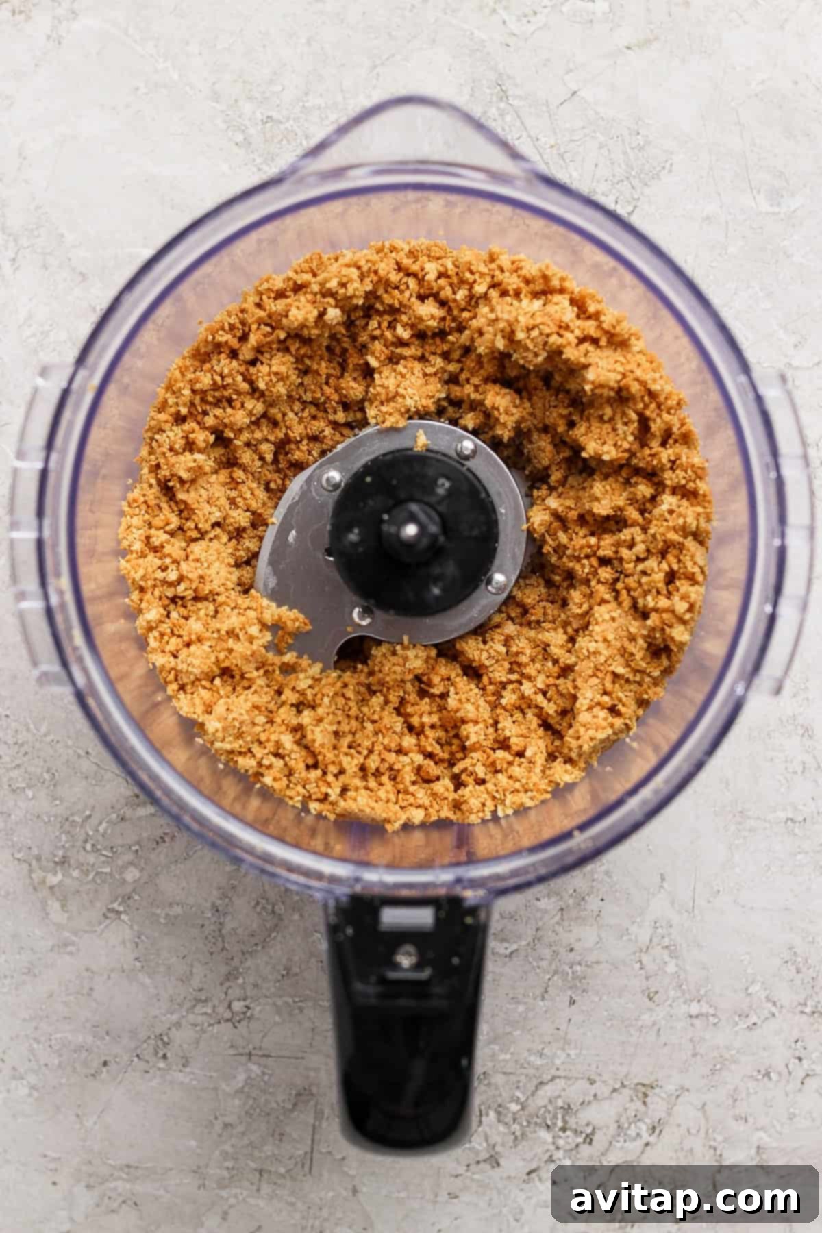 A food processor containing a mixture of finely ground graham crackers and butter, creating a crumbly crust base, on a light textured surface.
