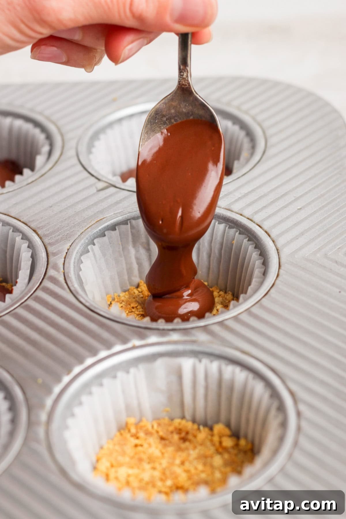 A hand holds a spoon pouring melted chocolate over crushed graham crackers in a paper-lined muffin tin, forming the crunchy layer.