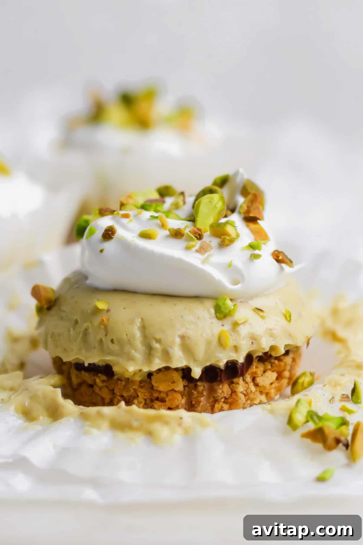 A perfectly portioned mini pistachio cheesecake with a crumb crust, layered filling, topped with a swirl of whipped cream and chopped pistachios, resting on white parchment paper.