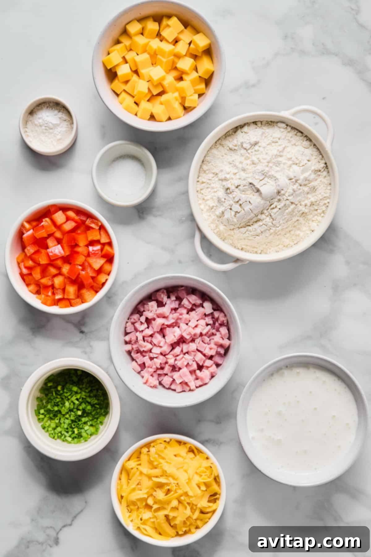 A collection of fresh, high-quality ingredients for making savory breakfast biscuits, arranged artfully on a marble countertop, including cubed cheddar, all-purpose flour, milk, diced ham, vibrant red bell pepper, shredded cheese, fresh chives, salt, and baking powder.