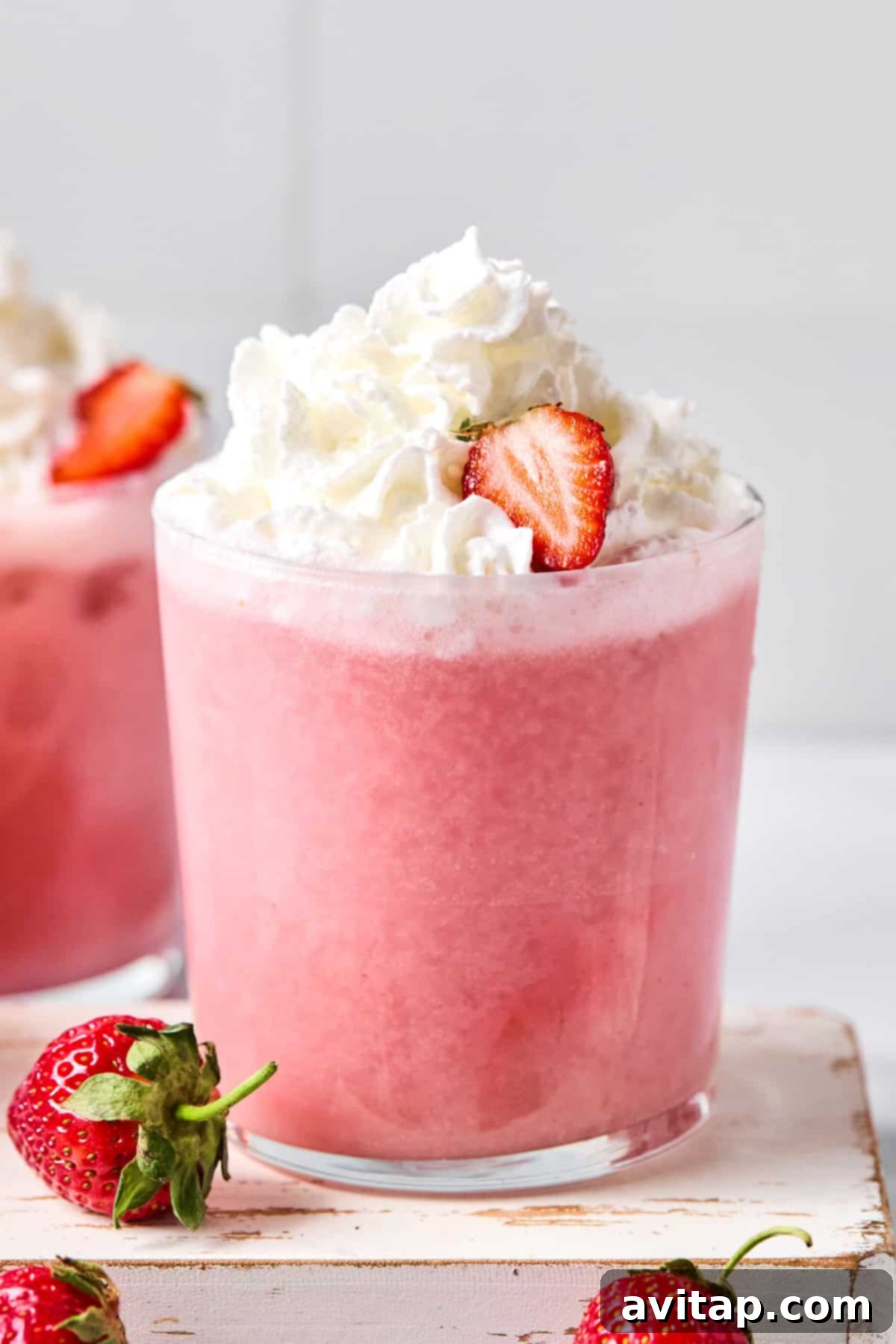 Quick Strawberry Dream Cocktail 2 A glass of pink strawberry smoothie topped with whipped cream and a sliced strawberry, with whole strawberries nearby.