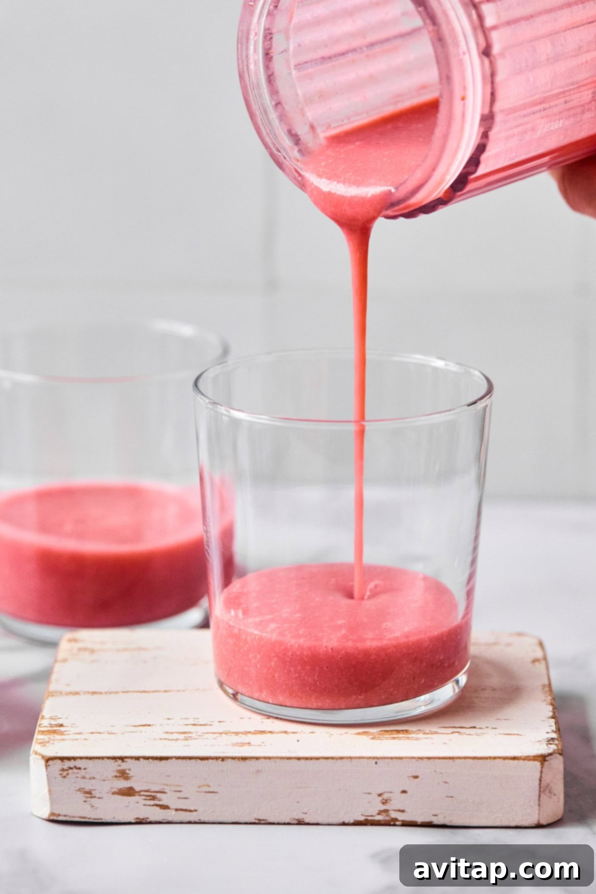 Quick Strawberry Dream Cocktail 3 Pink smoothie being poured from a blender into a glass, with another glass of smoothie nearby on a small white wooden board.