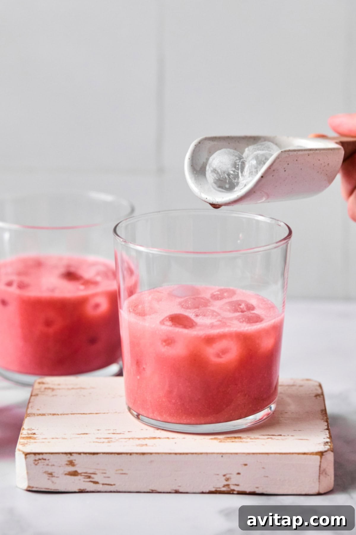Quick Strawberry Dream Cocktail 4 A hand adds ice cubes from a small white scoop into a glass of pink iced beverage, with another glass of the same drink in the background.