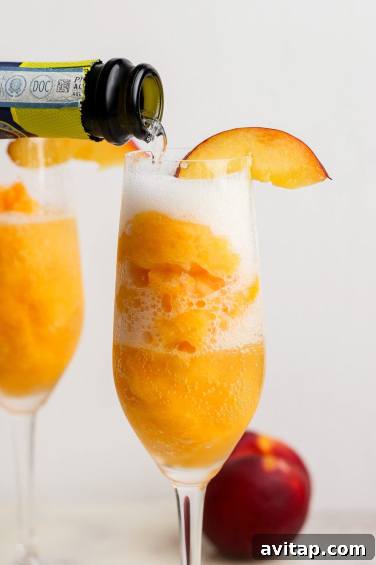 Luscious Frozen Peach Bellini 2 A glass filled with peach sorbet is being topped with sparkling wine, garnished with a peach slice; another glass and a peach are in the background.