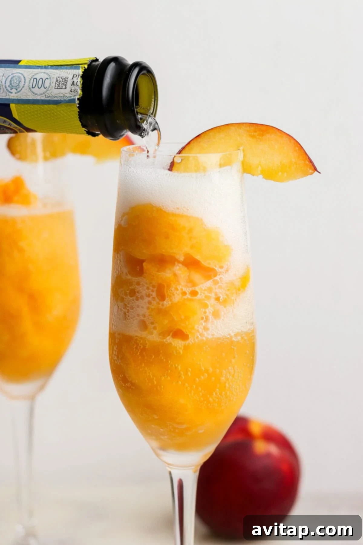 An elegant champagne flute, filled with vibrant peach sorbet, is being artfully topped with effervescent sparkling wine, garnished with a fresh peach slice. Another beautifully prepared bellini and whole peaches rest in the soft-focus background, ready to be enjoyed.