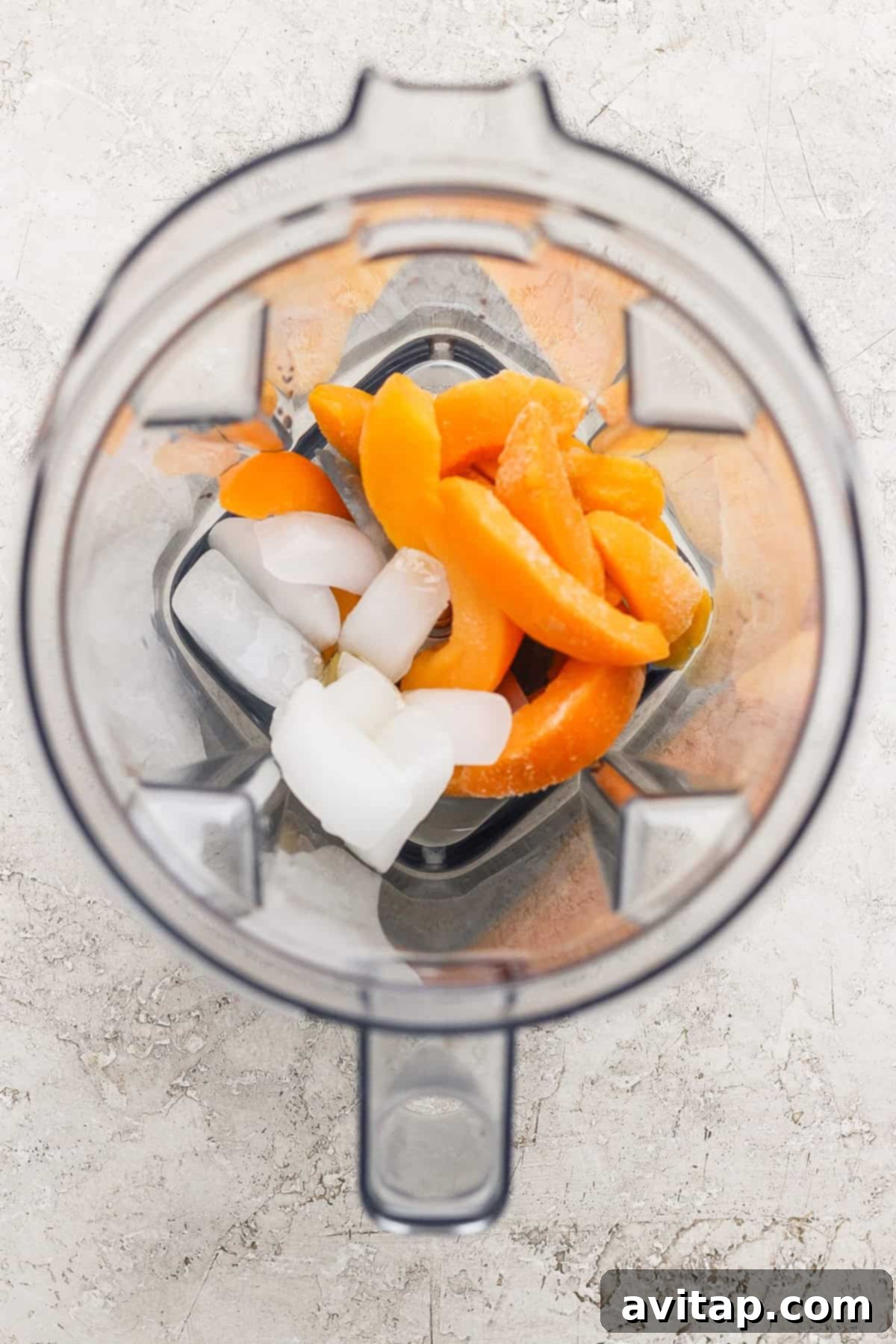 Luscious Frozen Peach Bellini 3 Top-down view of a blender containing ice cubes and orange fruit slices on a light-colored surface.