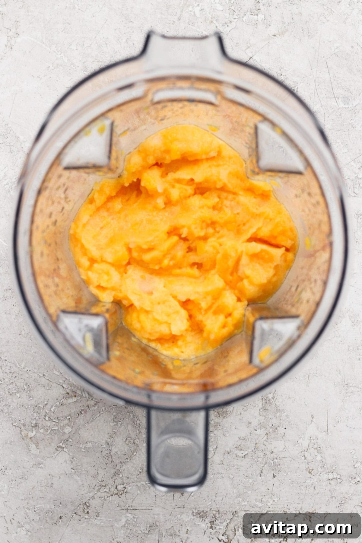 Luscious Frozen Peach Bellini 4 Top-down view of an open blender containing a smooth, orange-yellow mixture on a light textured surface.