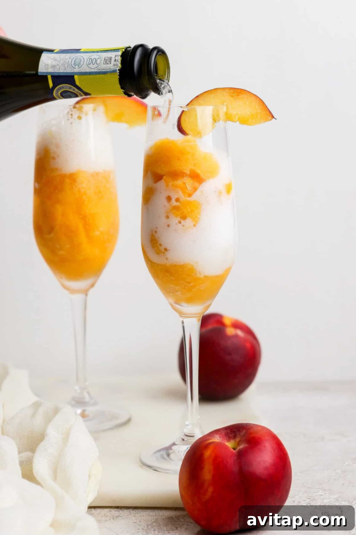 Luscious Frozen Peach Bellini 6 A glass of peach sorbet with sparkling wine being poured over it, garnished with a peach slice; another filled glass and whole peaches are in the background.