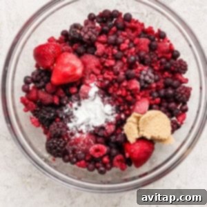 Creamy Protein Berry Cobbler 6 A glass bowl filled with mixed frozen berries, a mound of brown sugar, and a small amount of white powder on a light surface.