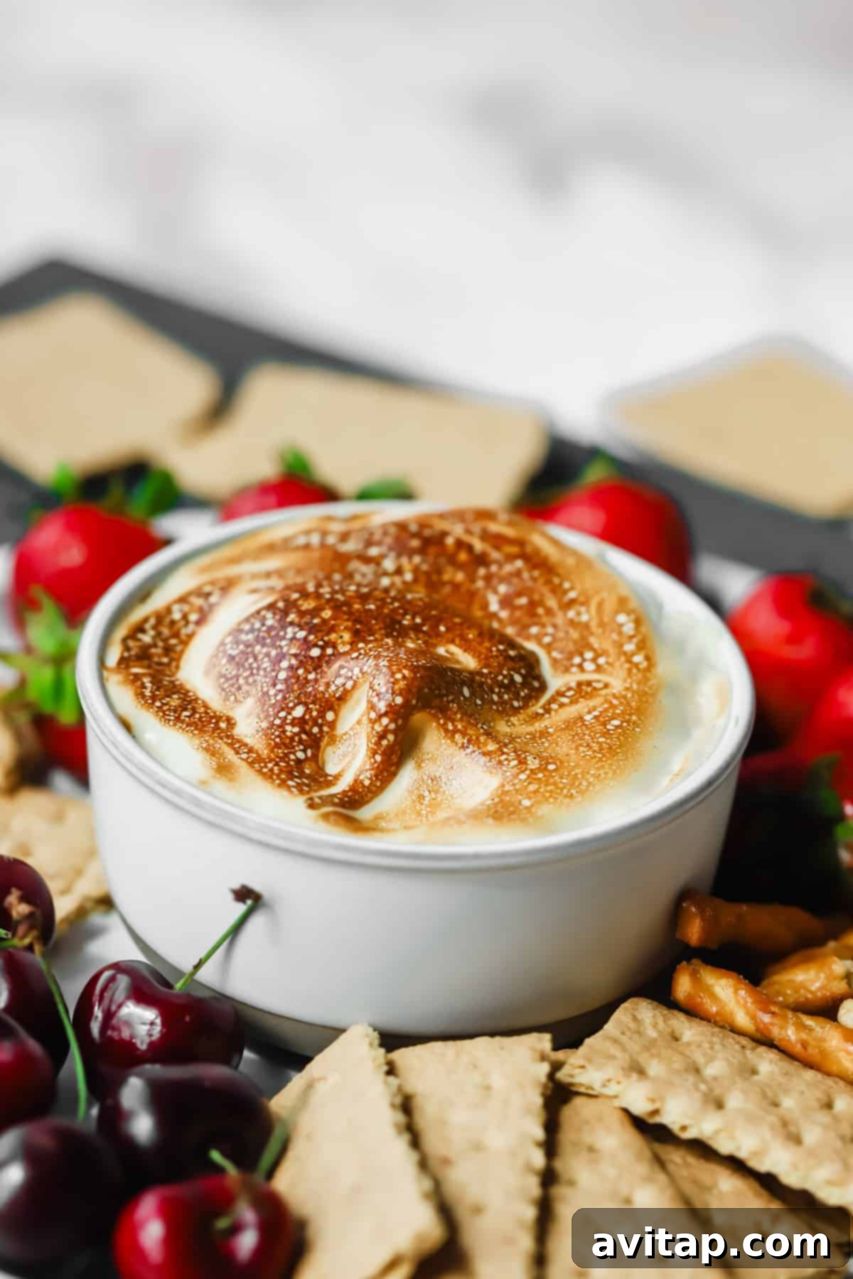 A white bowl filled with toasted maple marshmallow fluff, surrounded by a delightful array of graham crackers, crunchy pretzels, fresh strawberries, and vibrant cherries, inviting you to dip and enjoy.