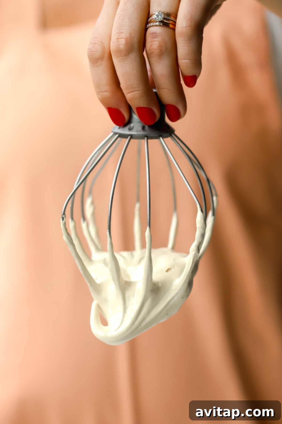 A hand with elegantly painted red nail polish holds a metal whisk, coated in thick, creamy maple marshmallow fluff, set against a soft, peach-colored background, showcasing the perfect texture.