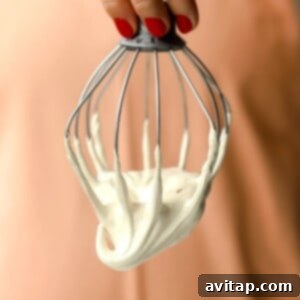 A hand with red nail polish holds a metal whisk covered in thick, creamy maple marshmallow fluff against a peach-colored background, highlighting its delightful texture.