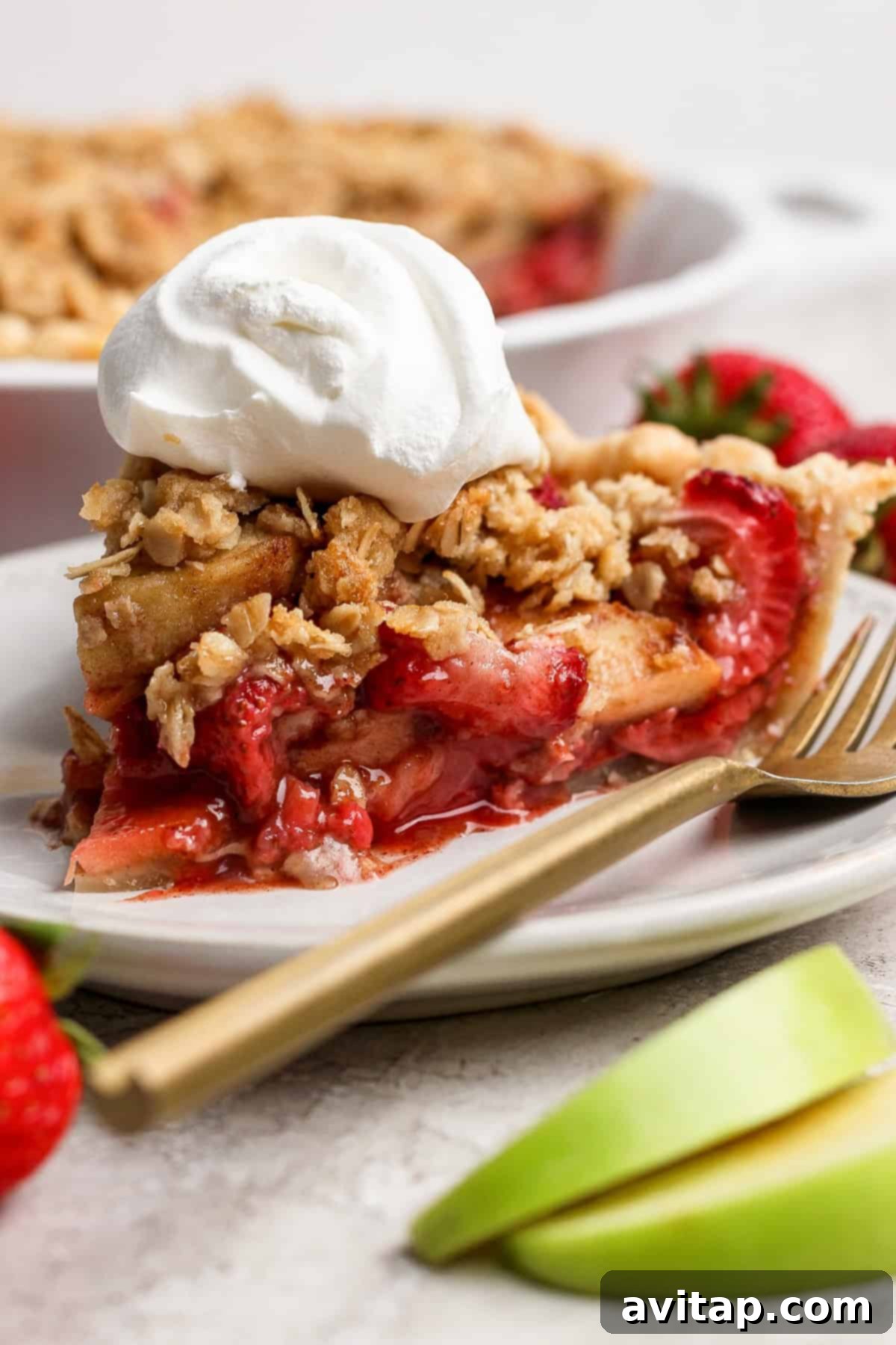 A generous slice of strawberry apple pie crumble, beautifully topped with a dollop of fresh whipped cream, rests on a pristine white plate. Alongside it, a golden fork, glistening fresh strawberries, and thinly sliced apples complete the inviting presentation, highlighting the pie's golden crust and rich filling.