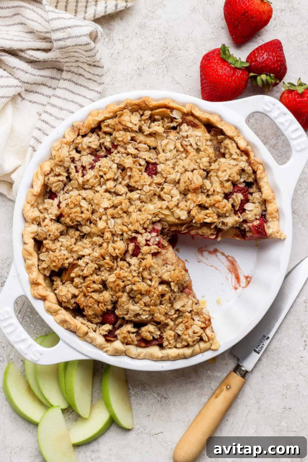 A beautifully baked strawberry apple pie with a golden oat crumble topping rests invitingly in a pristine white ceramic pie dish. A single, perfect slice has been neatly removed, revealing luscious layers of tender, spiced apples and sweet, vibrant strawberries within. Fresh strawberries, crisp apple slices, and a silver serving knife are artfully arranged beside the dish, enhancing the visual appeal of this rustic dessert.