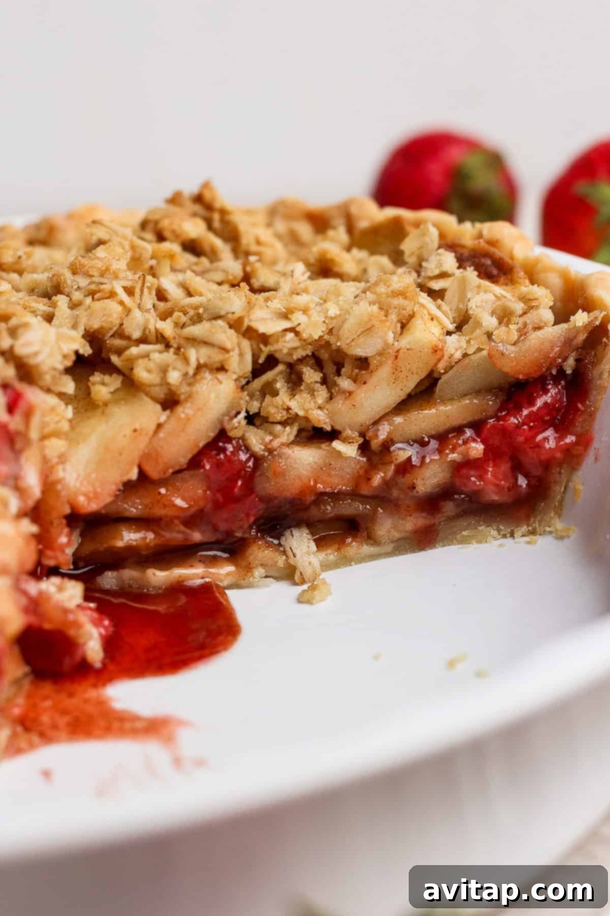 A mouth-watering close-up view of a sliced strawberry apple pie crumble. The golden oat topping is perfectly browned and inviting, and the interior reveals luscious, tender layers of spiced apples and sweet, vibrant strawberries, all beautifully nestled within a white ceramic pie dish, showcasing its rich texture and appealing colors.