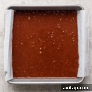 A rectangular metal baking pan lined with parchment paper is filled with s'mores brownies batter on a light textured surface.