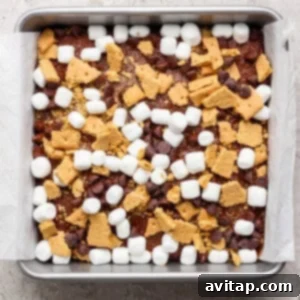 A square baking pan lined with parchment paper, filled with a mixture of mini marshmallows, chocolate chips, and broken graham crackers sits atop a rich s'mores brownies base.