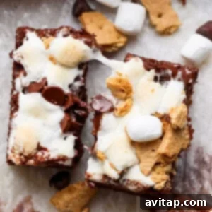 Two s'mores brownies topped with melted marshmallows, chocolate chips, and graham cracker pieces, placed on parchment paper with scattered ingredients around.