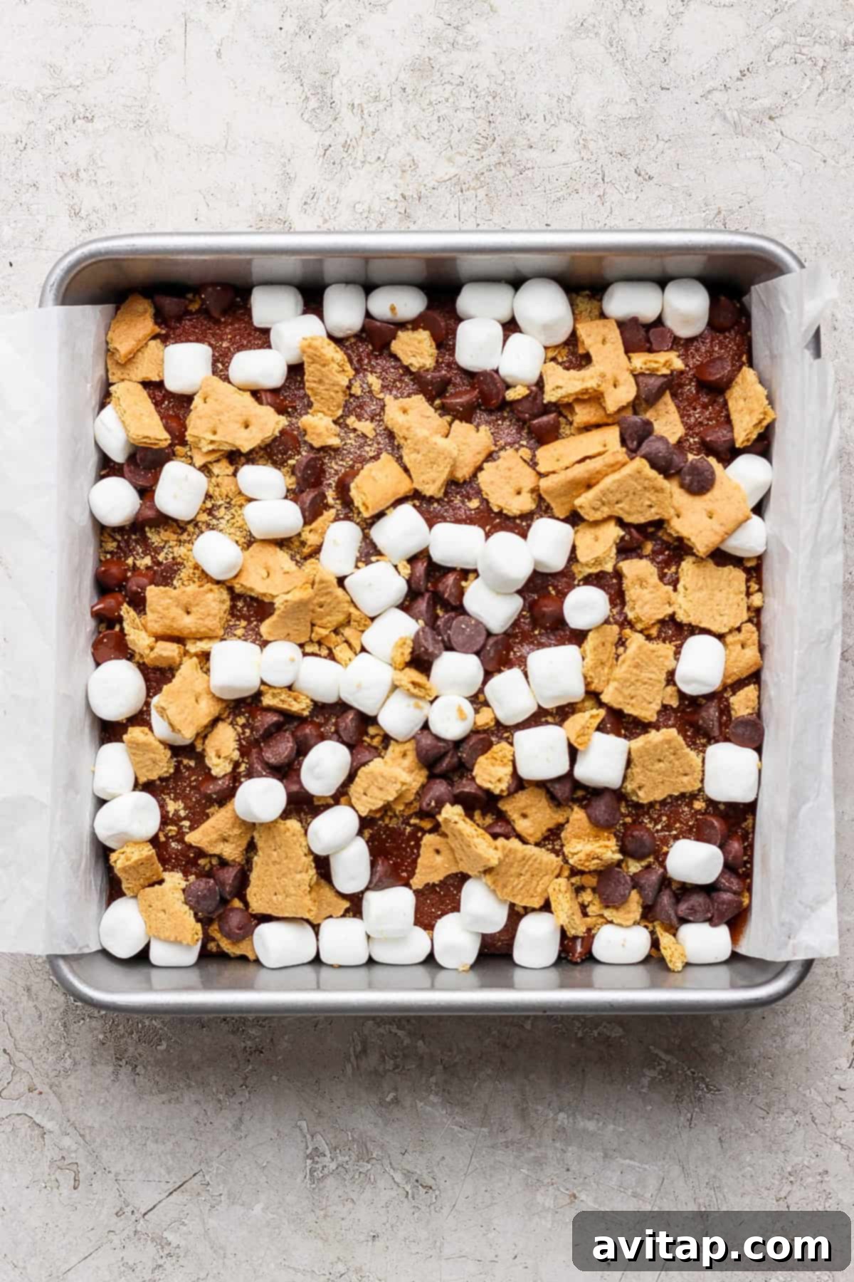 A square metal baking pan, lined with parchment paper, holds the rich s'mores brownie base generously topped with a mixture of mini marshmallows, chocolate chips, and broken graham cracker pieces, ready for baking.