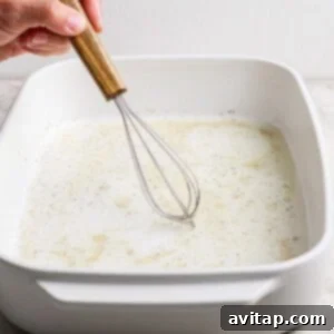 Grilled Rainbow Gnocchi Pops 7 A hand uses a wooden-handled whisk to mix a liquid with spices in a white rectangular baking dish on a light countertop.