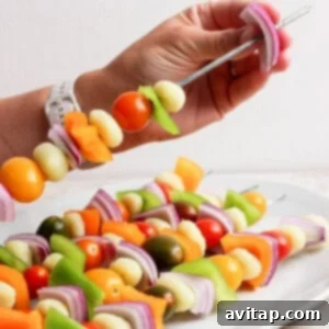 Grilled Rainbow Gnocchi Pops 8 A hand holds a metal skewer with pieces of onion, bell pepper, cherry tomatoes, and other vegetables above a plate of similar vegetable skewers.