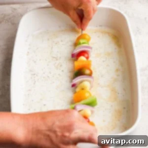 Grilled Rainbow Gnocchi Pops 9 A person places a skewer with vegetables—tomatoes, onions, peppers—into a white baking dish.