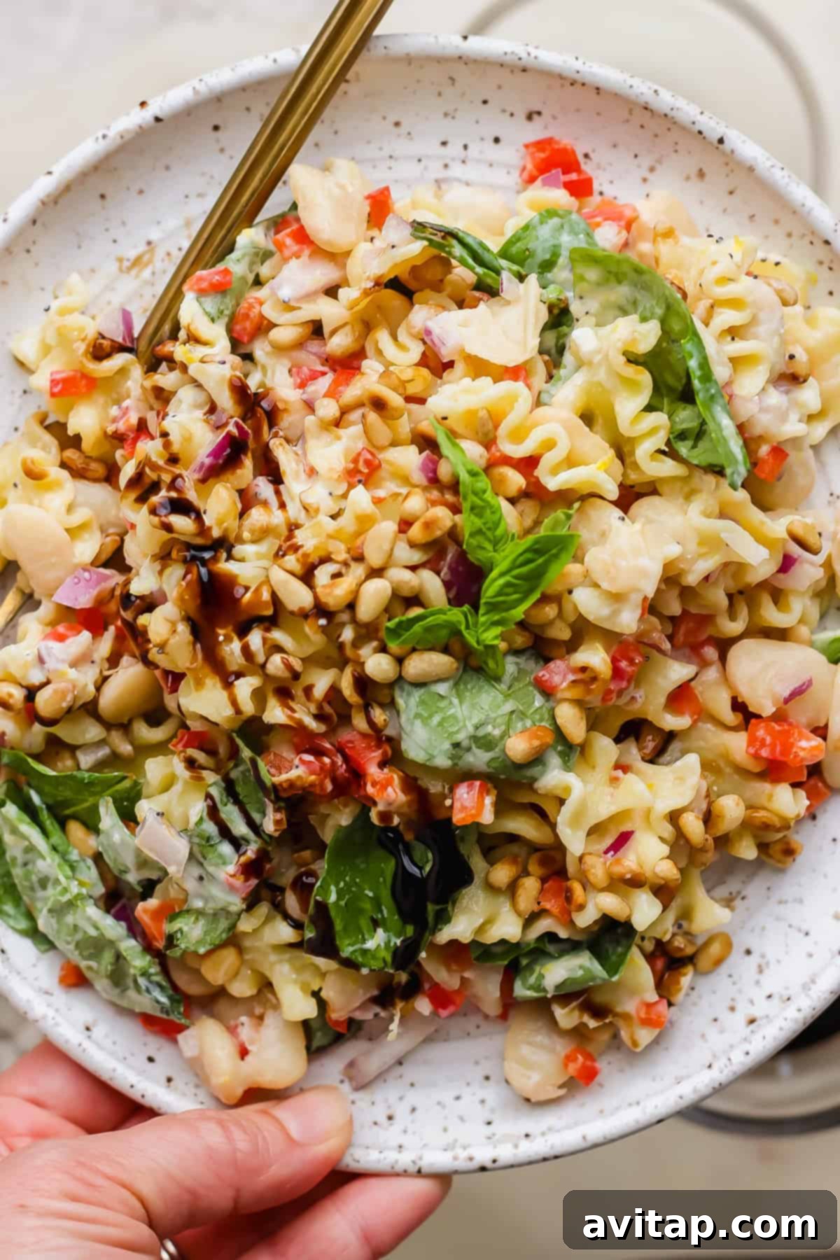 A plate of pasta salad with spinach, cannellini beans, red onion, diced red peppers, pine nuts, and a balsamic glaze, served with a fork.
