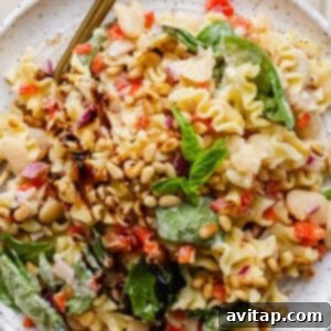 A plate of pasta salad with spinach, pine nuts, white beans, chopped red onions, red bell peppers, and a balsamic drizzle, garnished with fresh basil.