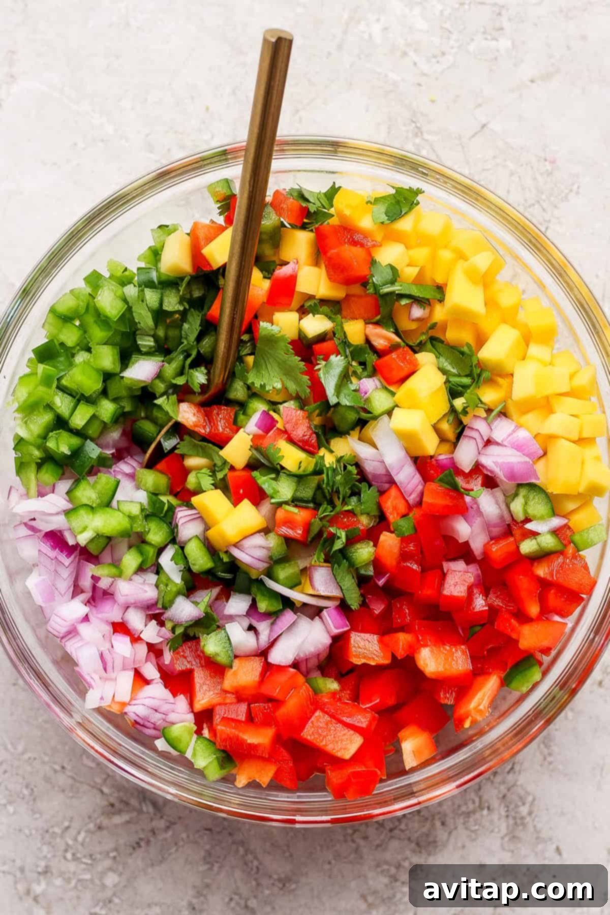 A clear glass bowl brimming with the colorful components of fresh mango salsa: finely chopped red onion, vibrant red and green bell pepper, juicy mango, spicy jalapeño, and fresh cilantro, with a gold serving spoon resting inside on a light surface. Ideal for healthy meal prep.