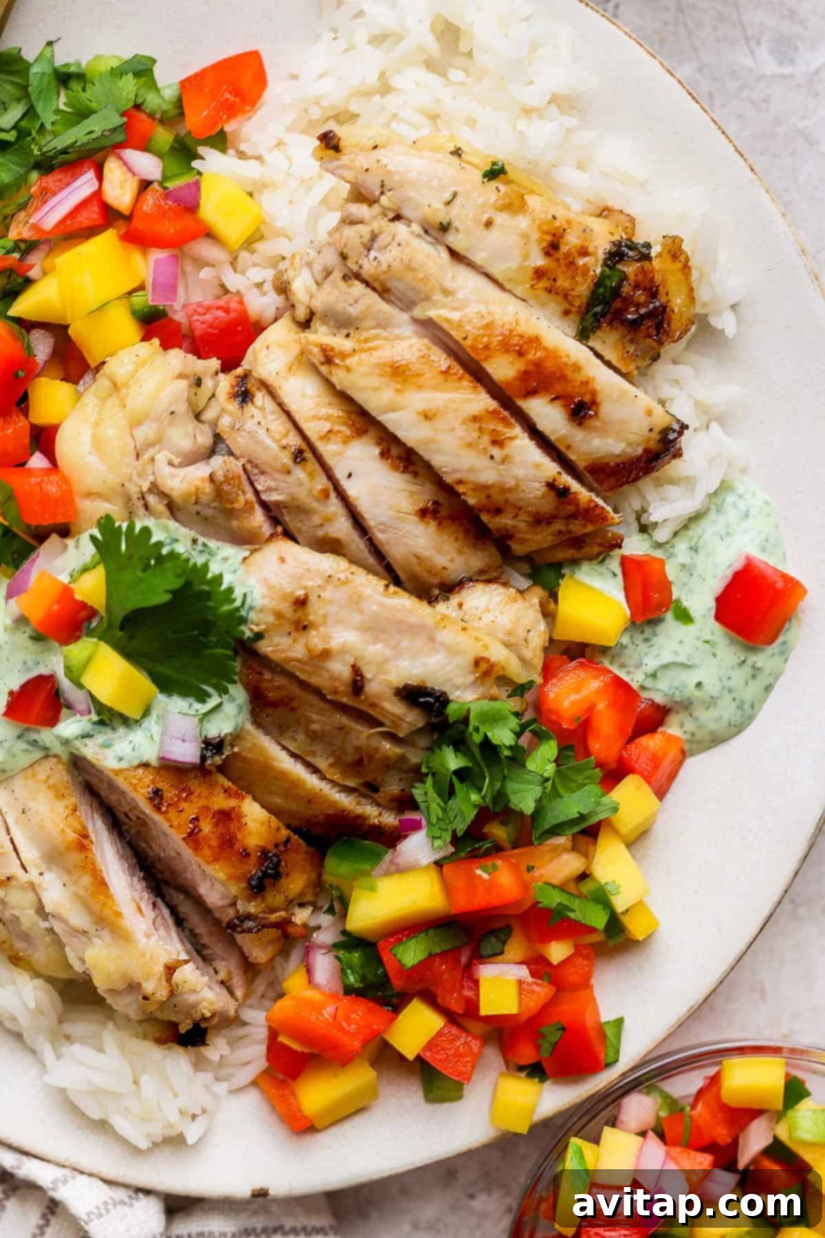 A perfectly prepared chicken meal prep plate featuring succulent grilled chicken slices artfully arranged on a bed of fluffy white rice, generously topped with a colorful diced mango salsa, garnished with fresh cilantro, and finished with a delicate drizzle of creamy herb sauce.