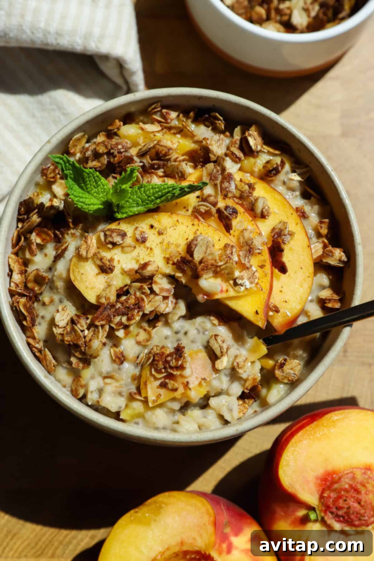 A bowl of oatmeal topped with granola, fresh peach slices, and a mint sprig, with a spoon inside and peach halves nearby on a wooden surface.