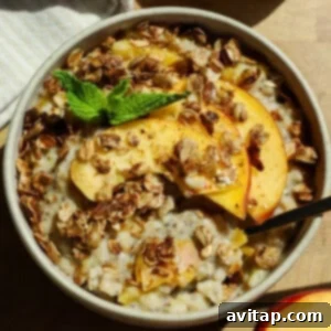 A bowl of oatmeal topped with granola, peach slices, and a sprig of mint, with a spoon placed inside.