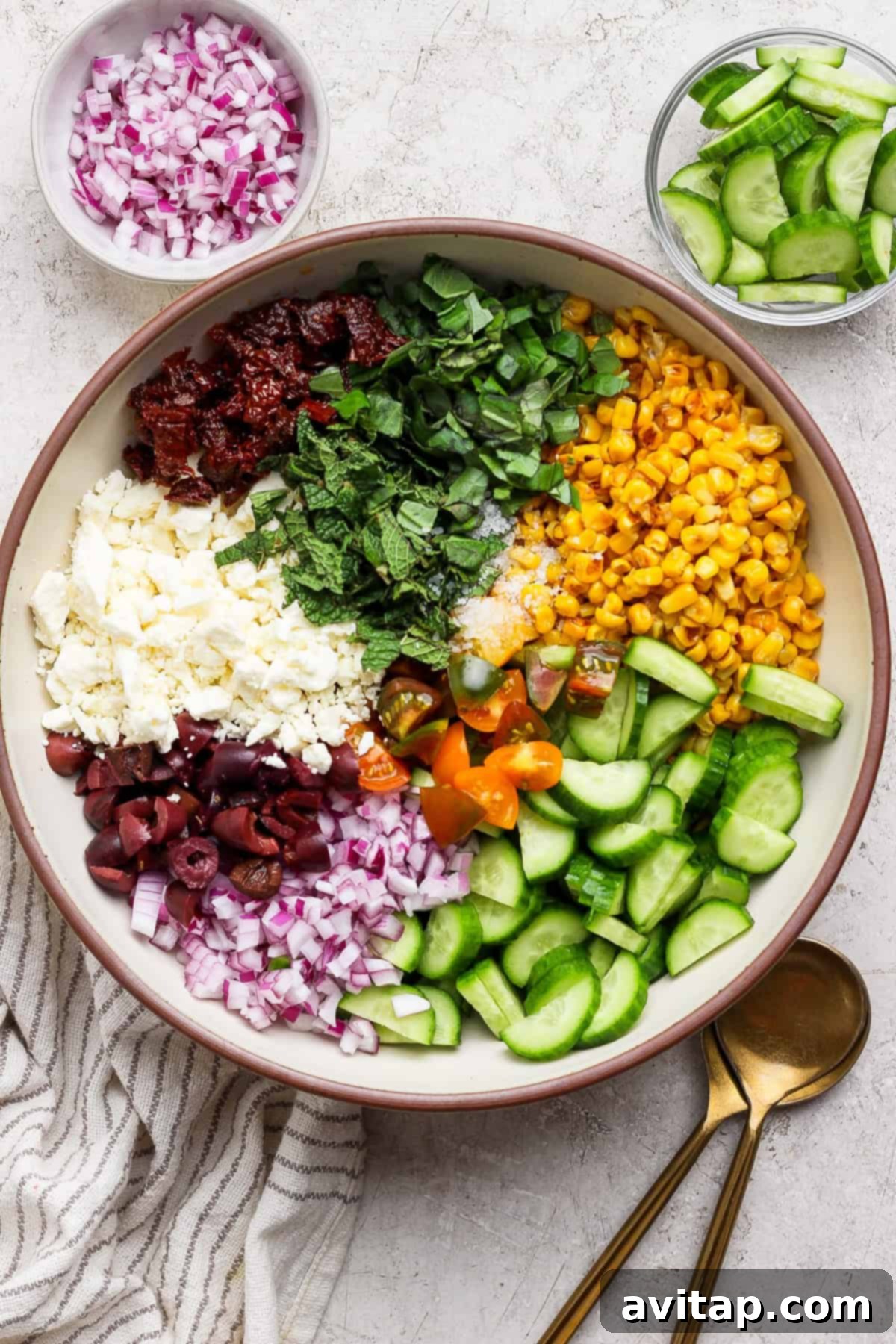 A large bowl containing chopped red onion, sun-dried tomatoes, feta cheese, olives, basil, mint, corn, cherry tomatoes, and cucumber, with bowls of onions and cucumbers on the side, ready to be mixed for a Mediterranean corn salad.