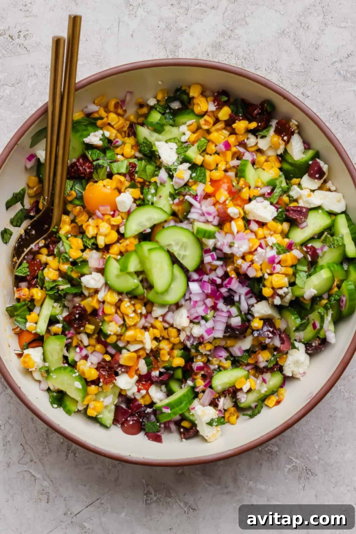 A bowl of Mediterranean corn salad with cucumbers, red onions, cherry tomatoes, feta cheese, leafy greens, and two gold forks, ready to be enjoyed.