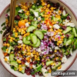 A bowl of chopped salad with corn, cucumbers, red onions, cherry tomatoes, feta cheese, and herbs, with a serving spoon, showcasing a vibrant Mediterranean corn salad.