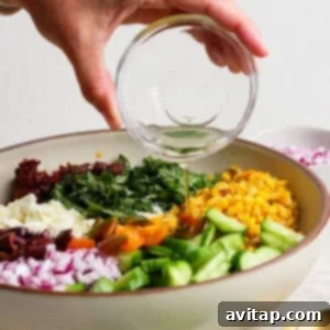 A hand pours dressing from a glass bowl onto a vibrant Mediterranean corn salad featuring cucumber, red onion, tomatoes, olives, feta cheese, and greens in a large white bowl.