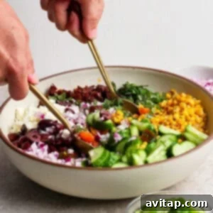 A person prepares a mediterranean corn salad, mixing chopped cucumber, corn, red onion, olives, sun-dried tomatoes, greens, and feta cheese in a large bowl using two gold utensils.